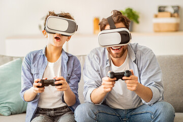Virtual reality. Excited father and teen son trying VR glasses and playing videogames together, holding joysticks, sittin gon sofa at home interior. Leisure time with dad