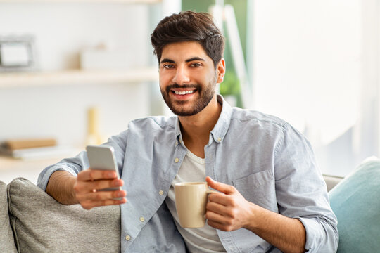 Morning routine. Modern arab guy checking emails and news on smartphone and drinking coffee. Man scrolling feed on phone, sitting on comfortable sofa at home and smiling to camera