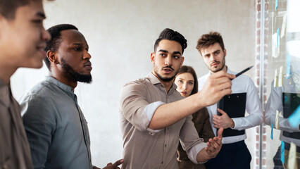 Diverse group of young serious multiethnic business people having meeting, using sticky paper post-it notes on glass wall or window to share ideas and thinking. Brainstorming And Workshop Concept