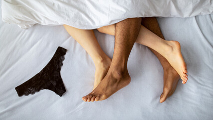 Top view of young multiracial couple making love on bed, cute panties lying nearby. Overhead shot of passionate millennial lovers having sex, playing erotic games, engaged in sexual foreplay