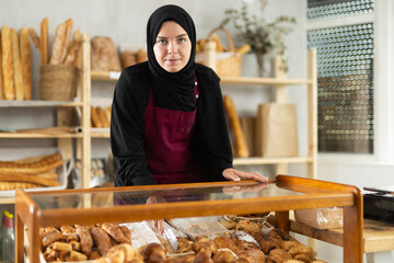 Muslim girl seller in hijab stands at bakery counter with basket filled with fresh croissants....