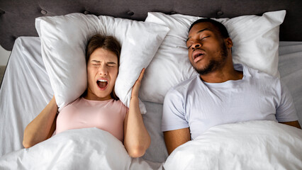 Top view of snoring black husband and his stressed wife screaming and covering ears with pillow in bed at home. Millennial lady cannot sleep, being disturbed by loud noise, shouting in anger