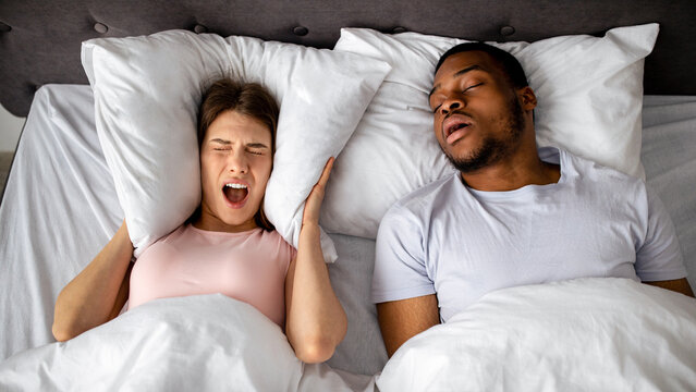 Top view of snoring black husband and his stressed wife screaming and covering ears with pillow in bed at home. Millennial lady cannot sleep, being disturbed by loud noise, shouting in anger