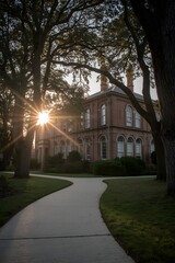 Historic red brick building with arched windows at sunrise framed by large trees and manicured garden path