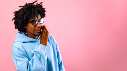 Portrait of black teenage guy whispering secret, sharing confidential information, spreading gossip on pink studio background. African American teenager communicating rumor or private info