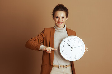 A smiling businesswoman in a blazer points to a large wall clock, addressing the viewer. It conveys...