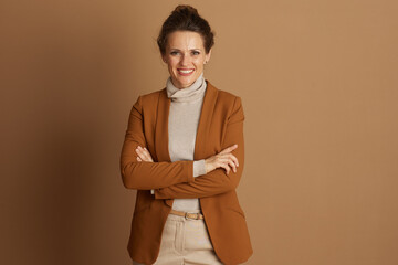 Obraz premium A confident businesswoman with bun hairstyle smiles at the camera, arms crossed. Wearing brown blazer and beige turtleneck, she exudes professionalism and approachability against warm brown background