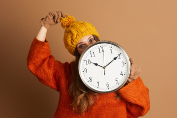 A playful woman in an orange sweater, yellow beanie, and brown gloves peeks over a wall clock with...