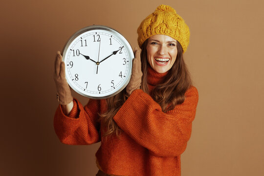 A smiling young woman in a yellow beanie and orange turtleneck sweater holds a large wall clock against a light brown background. The scene conveys a cheerful, cozy, and seasonal mood.