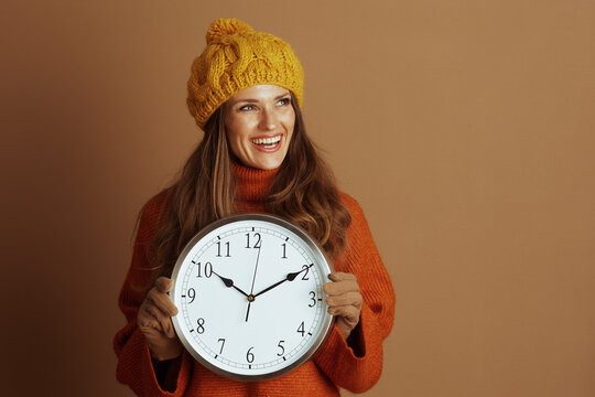 A smiling woman in a warm orange turtleneck and yellow beanie happily holds a large analog clock, looking to her right. The scene conveys a cheerful, optimistic mood about seasonal changes.