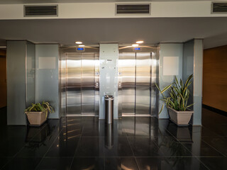 Two steel lift doors in a modern office lobby, with floor indicators, call buttons, glass panels...