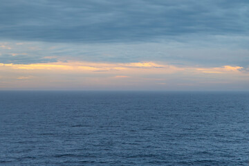 Vast ocean expanse under a cloudy sky at dawn, with a hint of golden sunlight on the horizon.
