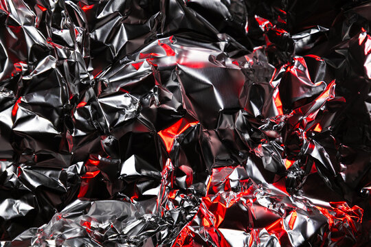 Close up of crumpled aluminum foil with shiny silver surface and dramatic red highlights. Abstract metallic background with reflective texture and artistic light play.