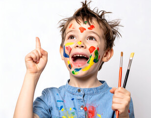 Joyful boy covered in colorful paint, holding brushes, pointing up with an idea. Young artist's creative moment and bright expression.