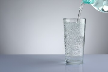 Pouring refreshing soda water from bottle into glass at white mirror table against light grey gradient background, closeup. Space for text