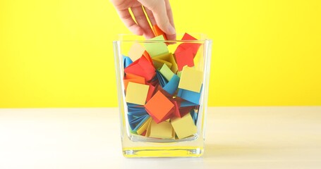Woman picking orange paper piece from glass jar at wooden table against yellow background, closeup