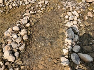A close-up aerial view of a dry riverbed with smooth, rounded stones and a sandy center, showcasing the natural texture and patterns of the earth