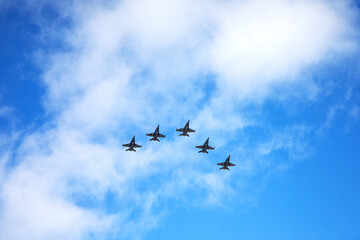 Five fighter jets soaring in perfect formation across a partly cloudy blue sky.
