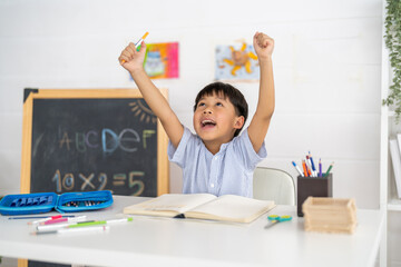 Excited young asian boy celebrating success while studying at home, raising hands in joy with open notebook, creative classroom, education and happy school moment, celebrating academic achievement