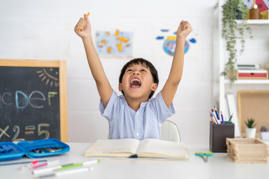 Excited young asian boy celebrating success while studying at home, raising hands in joy with open notebook, creative classroom, education and happy school moment, celebrating academic achievement - Powered by Adobe