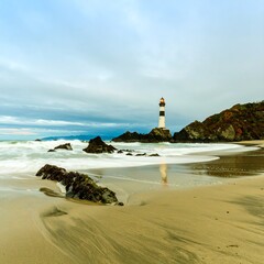 Lighthouse on Rocky Shoreline with Crashing Waves and Sandy Beach.