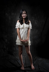 Naklejka premium Young Asian woman standing and smiling in studio, wearing off-shoulder white blouse and layered skirt, posing gracefully with one hand on chest and one leg raised, dark background.