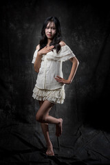 Naklejka premium Young Asian woman standing and smiling in studio, wearing off-shoulder white blouse and layered skirt, posing gracefully with one hand on chest and one leg raised, dark background.