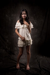 Fototapeta premium Young Asian woman standing and smiling in studio, wearing off-shoulder white blouse and layered skirt, posing gracefully with one hand on chest and one leg raised, dark background.