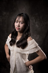 Young Asian woman standing and smiling in studio, wearing off-shoulder white blouse and layered skirt, posing gracefully with one hand on chest and one leg raised, dark background.