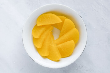 An overhead shot of a white plate filled with sliced yellow pickled radish (danmuji), arranged in a fan-like pattern on a marble background.