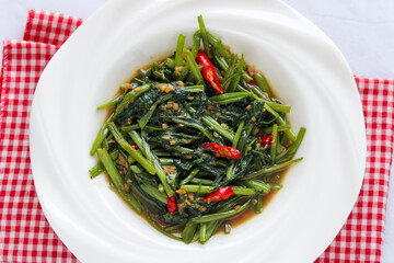 A closeup, overhead shot of a white bowl filled with stir-fried water spinach (kangkung) and red chili slices in a savory brown sauce.