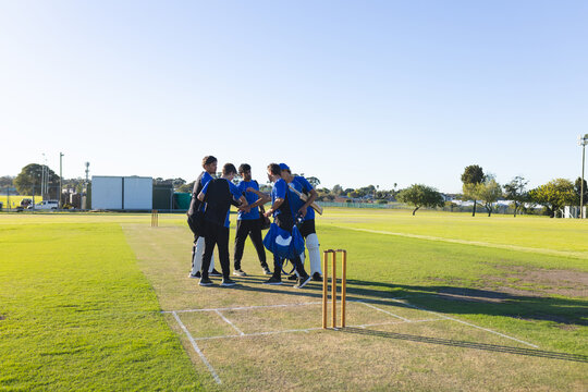 Six cricket players in blue jerseys holding bats in huddle on green field near wicket stumps