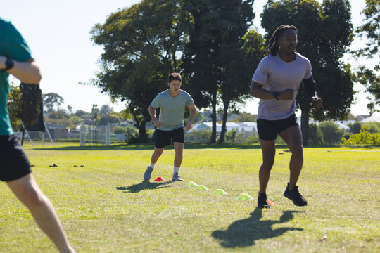 Diverse male athletes running between plastic agility cones on green grass field, copy space