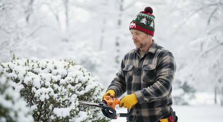 Mature landscaper wearing christmas knitted beanie using chainsaw trimming snow covered bushes in winter garden, seasonal tree care maintenance service