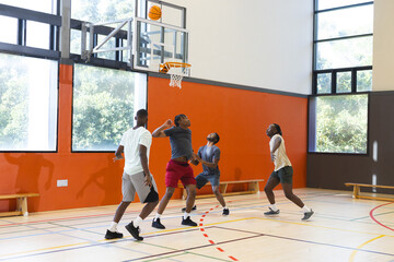 African american male friends playing basketball on gym court with hoop, benches