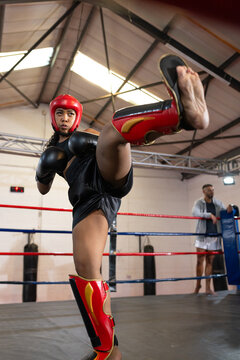 Female athlete executing high front kick in boxing ring in red headgear with trainer observing