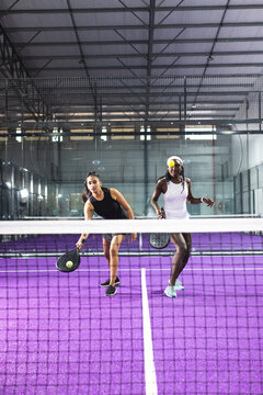 Diverse female teammates using rackets hitting yellow-green ball over net on padel court