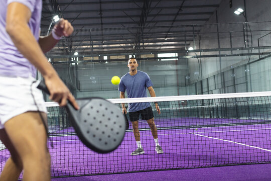 Two men playing padel tennis on purple court inside sports facility with rackets, yellow ball