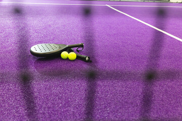 Black padel racket lying next to two tennis balls on purple court behind blurred net