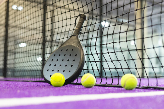 Black padel racket is leaning against net on purple turf court, yellow balls aligned next to line