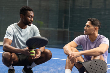 South asian and african american men crouching on padel court holding rackets, ball