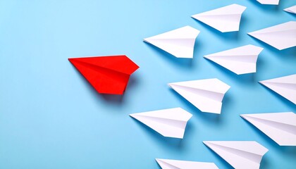 A red paper airplane stands out from a group of white paper airplanes on a light blue surface