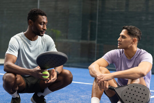 South asian and african american men crouching on padel court holding rackets, ball