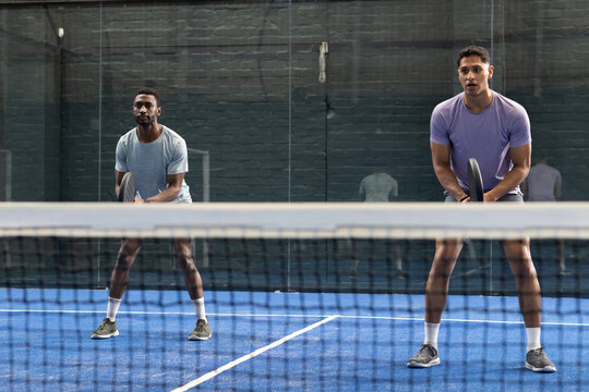 Diverse male partners gripping padel rackets, practicing low stance behind net on synthetic court