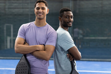 Diverse male teammates posing back-to-back on padel court with net holding padel rackets