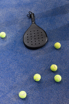 Black padel racket lying on blue textured court surface among yellow tennis balls