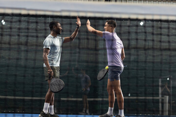 Diverse male teammates giving high-five on blue padel court with net, padel rackets, yellow ball