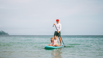 Children laugh and splash water while father paddles SUP board on sea. Happy playtime, joyful bonding, and healthy recreation with family on warm sunny day.