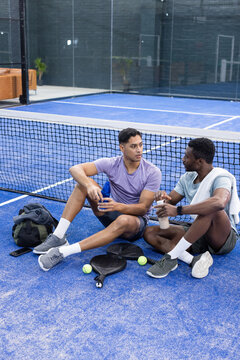 Diverse male friends resting on blue padel court turf beside water bottles rackets, balls