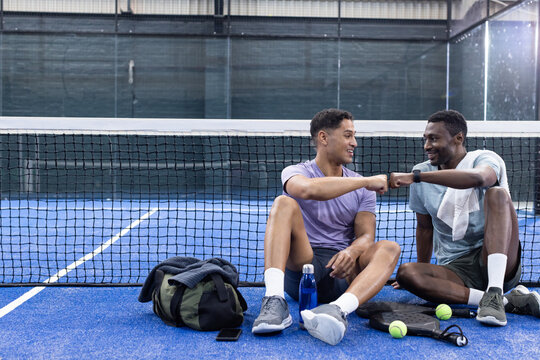 Diverse male friends sitting on blue padel court exchanging fist bump by green bag, racket